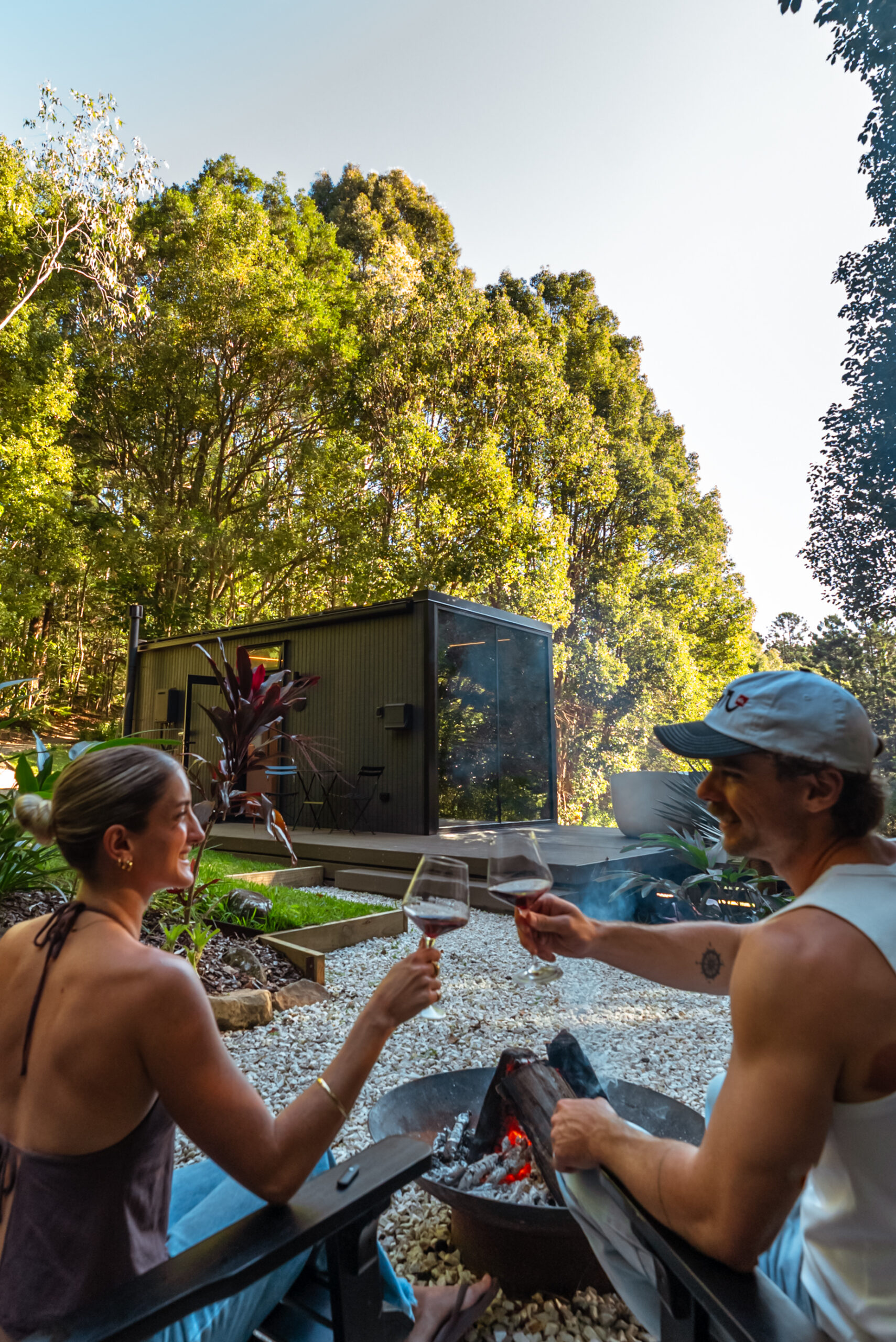 Couple toasting with red wine beside a firepit in front of the DSTNTN Mirror Home, set among tall rainforest trees in the Byron Bay