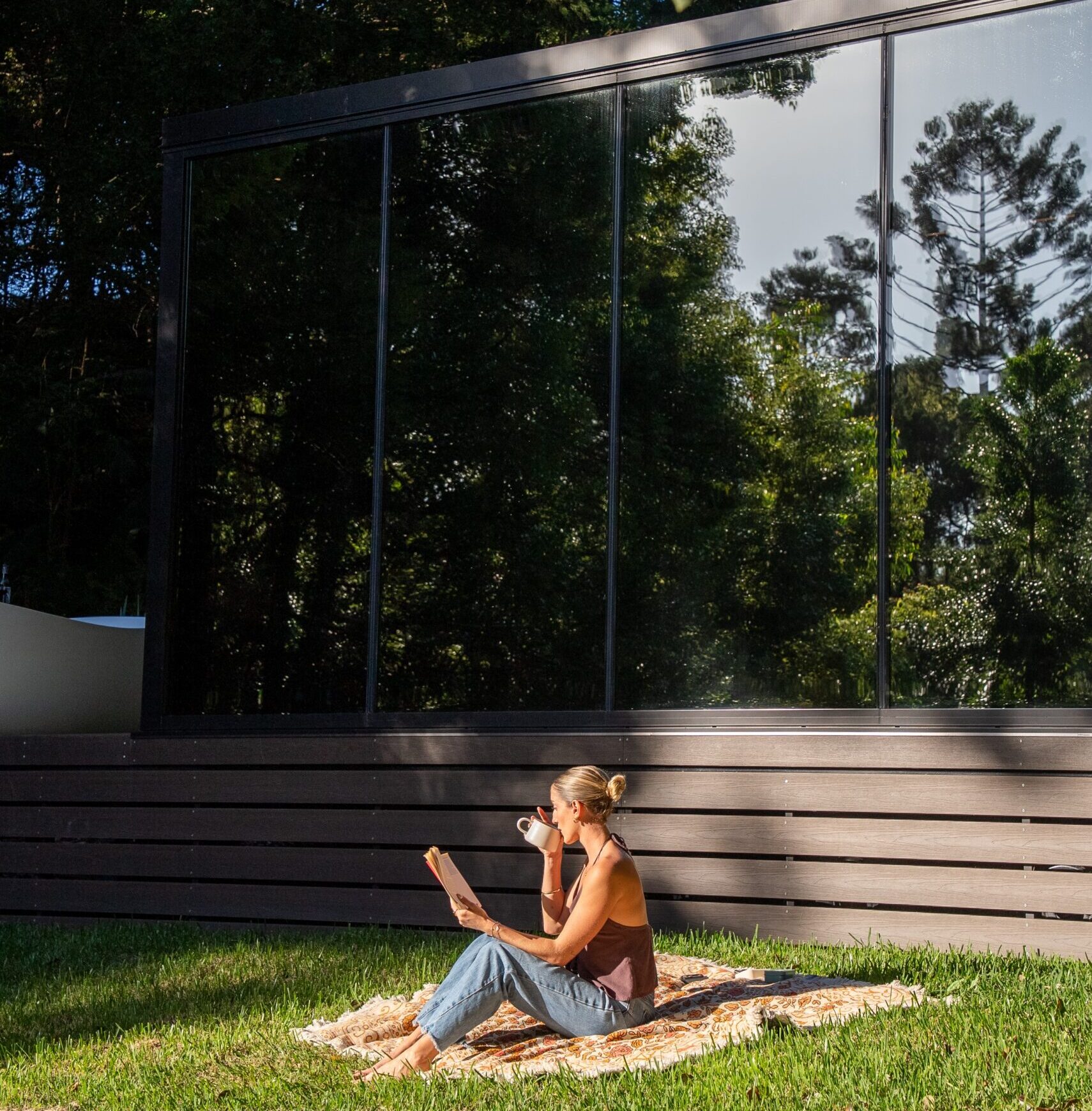 The science of novelty in action: a woman reads and sips coffee on a picnic rug in front of a DSTNTN Mirror Home, its glass reflecting the surrounding rainforest