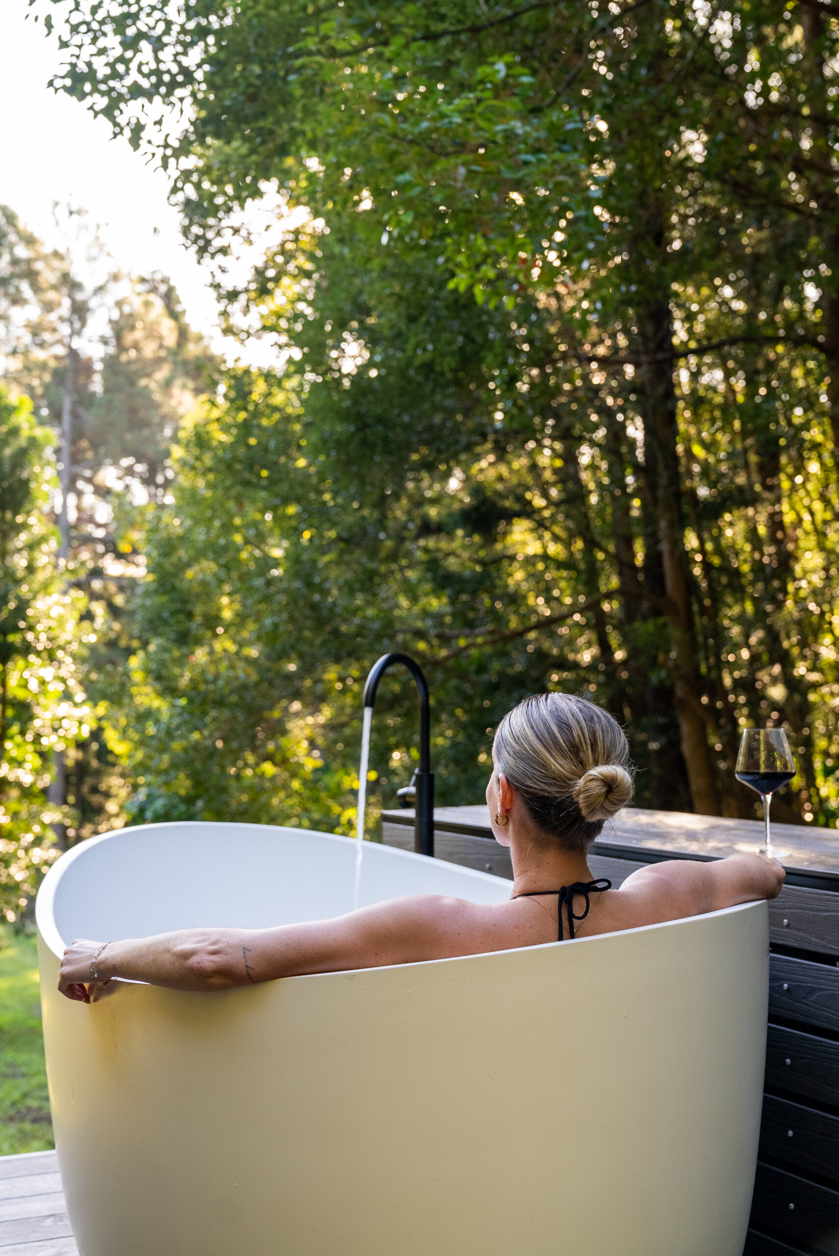 Woman relaxing in a freestanding outdoor bath with a glass of red wine, surrounded by rainforest at DSTNTN Byron Bay