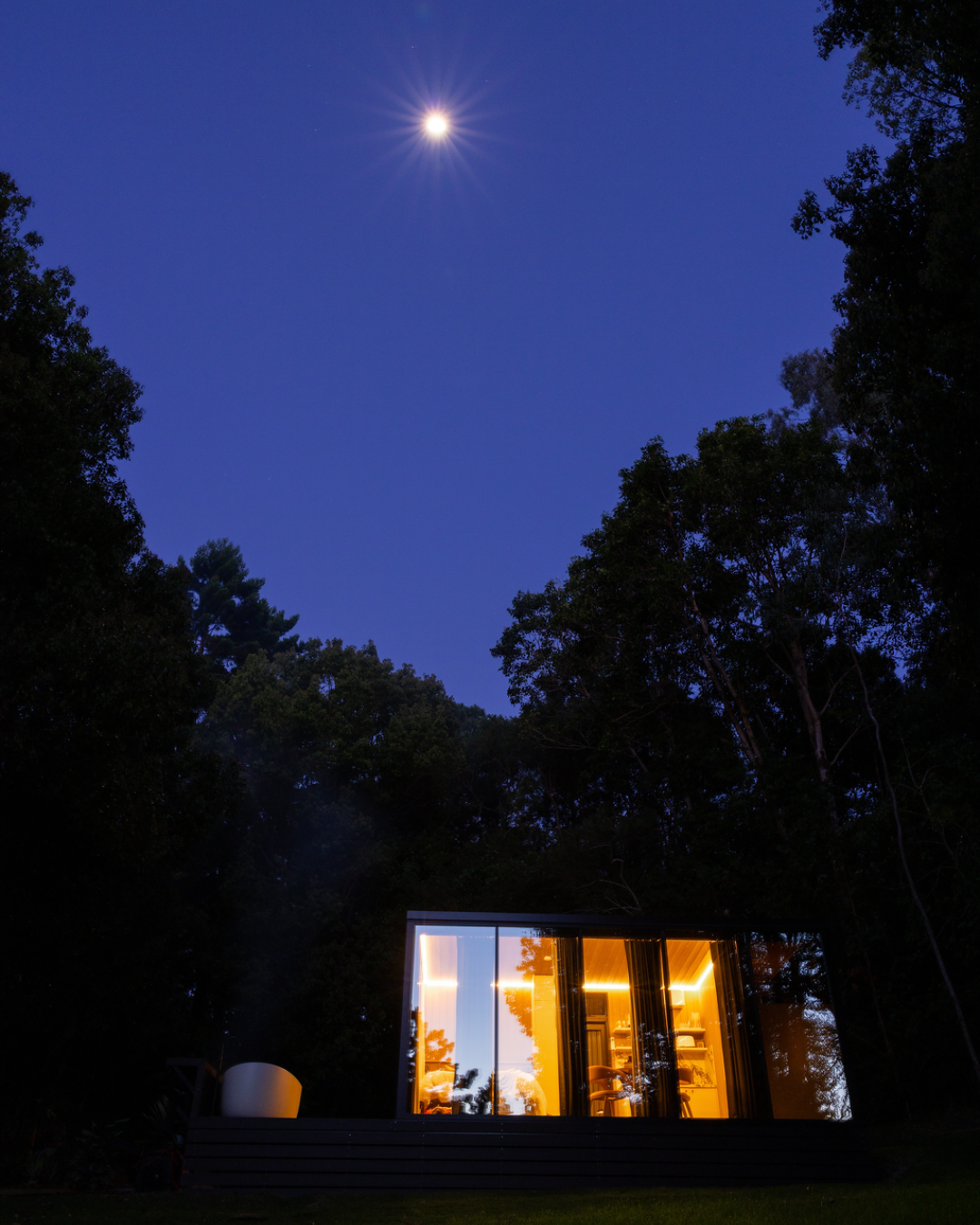 A DSTNTN Mirror Home glowing with warm light at dusk, framed by tall rainforest trees under a deep blue sky and a bright full moon